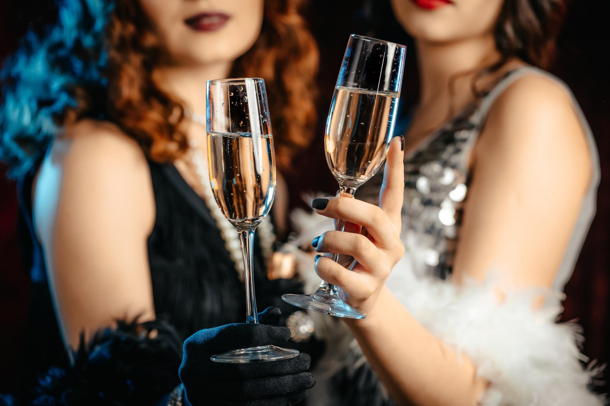 Close up glasses of champagne. Flappers women wearing in style of Roaring Gatsby twenties drinking alcohol. Vintage, retro party, fashion, girls friends concept.