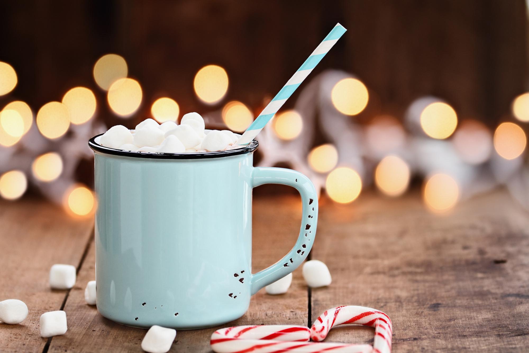Enamel cup of hot cocoa with mini marshmallows and candy canes in the shape of a heart against a rustic background with beautiful Christmas lights of bokeh. Could also be coffee. Perfect winter time treat.