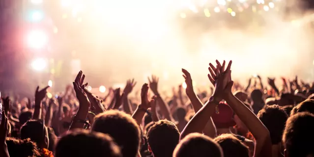 Audience with hands raised at a music festival and lights streaming down from above the stage. Soft focus, blurred movement..
