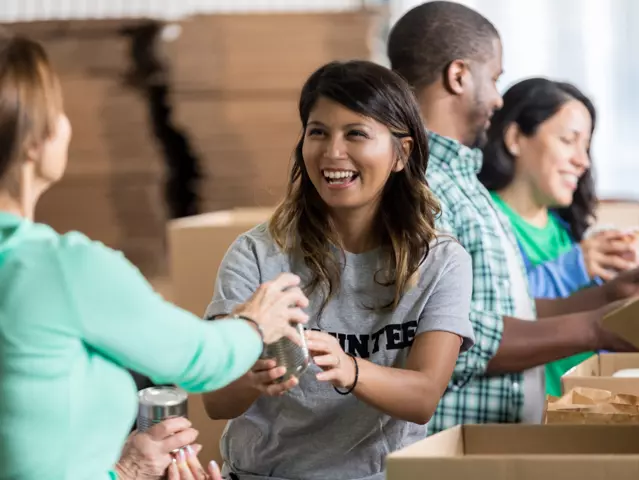 Cheerful mid adult Hispanic female volunteer accepts a canned food donation during community food drive. She is wearing a gray volunteer t-shirt. Her friends are volunteering in the background.