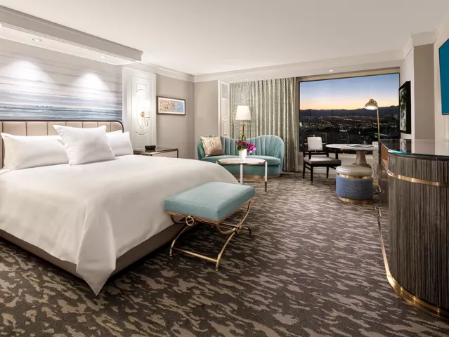 Bellagio hotel bedroom with a large bed dressed in crisp white linens, a padded headboard, modern wood furnishings, and a window overlooking the Las Vegas skyline.