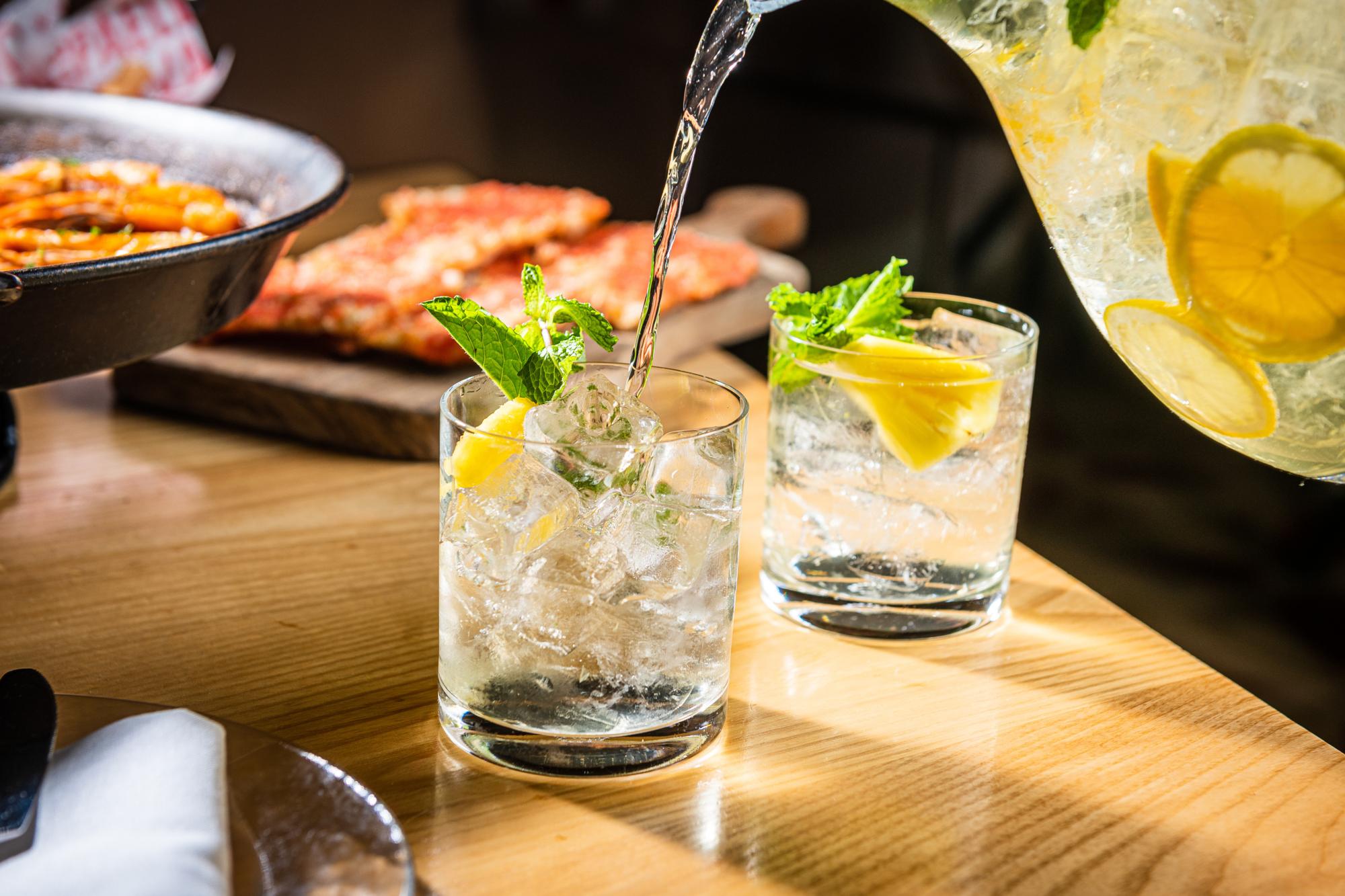 Bartender pouring sparkling beverage into glass of Cava Sangria with citrus and ice on wooden table.