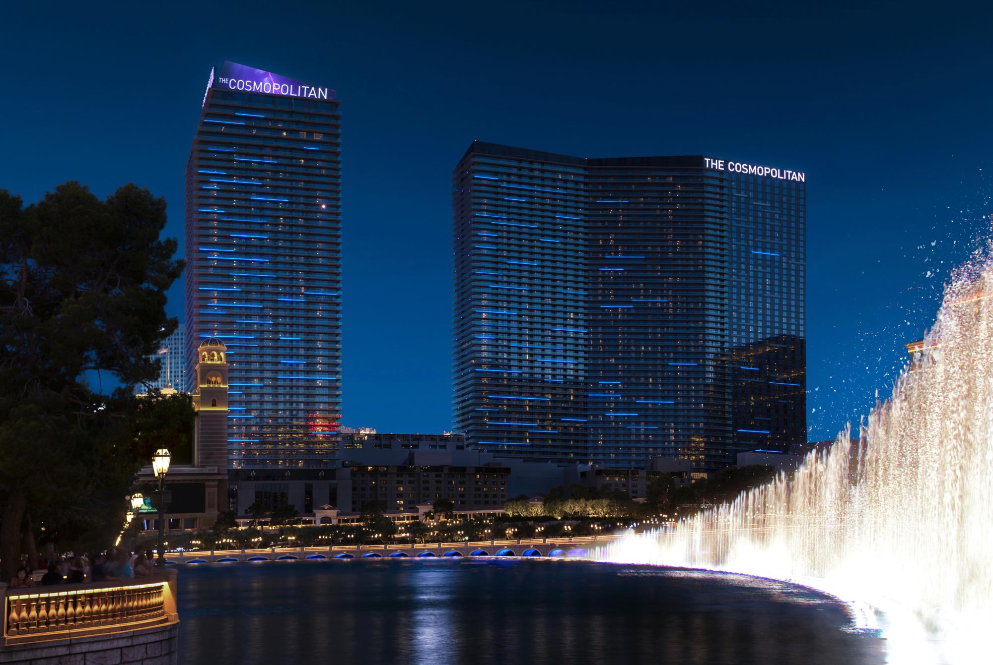 Exterior of The Cosmopolitan of Las Vegas at night shows two buildings with 40 floors each in front a lake with fountains.