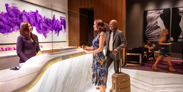 Woman and man with a suitcase at the reception of ARIA are attended by a receptionist at a white marble desk.