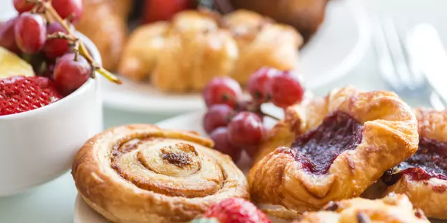 Closeup of a continental breakfast at a hotel, with bowls and plates filled with a selection of breakfast bakery items and fresh fruit on a white table with cutlery in the background.