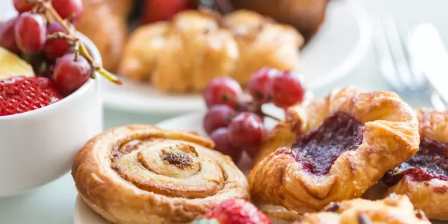 Closeup of a continental breakfast at a hotel, with bowls and plates filled with a selection of breakfast bakery items and fresh fruit on a white table with cutlery in the background.