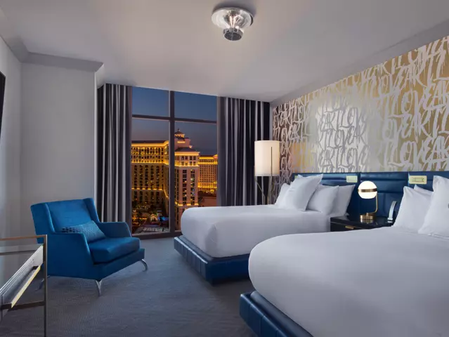 The Cosmopolitan hotel bedroom with a two large beds dressed in crisp white linens, modern wallpaper, and a window overlooking the Las Vegas skyline.