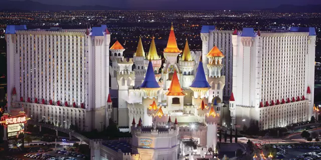 Exterior of Excalibur shows two large white buildings with 28 floors and 11 castle-like towers in the between the two buildings with a sunset mountain view.