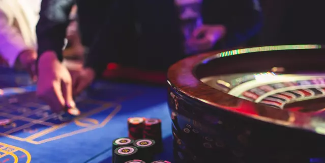 Vibrant casino table with roulette in motion, with casino chips, tokens, the hand of croupier, dollar bill money and a group of gambling rich wealthy people playing bet in the background"n