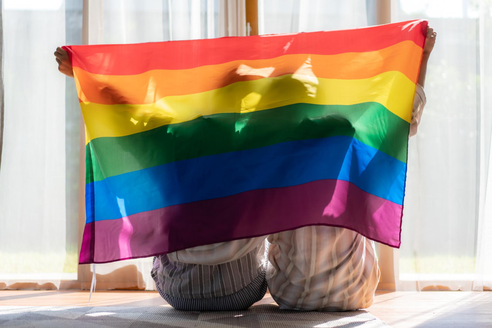 Asian couple Gay celebrating Pride LGBTQI at home on their own, with their partner with roommates and covering body by rainbow flag. Asian homosexsual man celebrating together with pride flag in home.